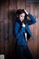 A woman in a denim jacket leaning against a wooden wall.