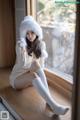 A woman sitting on a window sill wearing a white sweater and a furry hat.