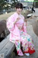 A woman in a pink kimono sitting on a bench next to a deer.