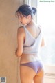 A woman in a gray top and purple panties leaning against a window.